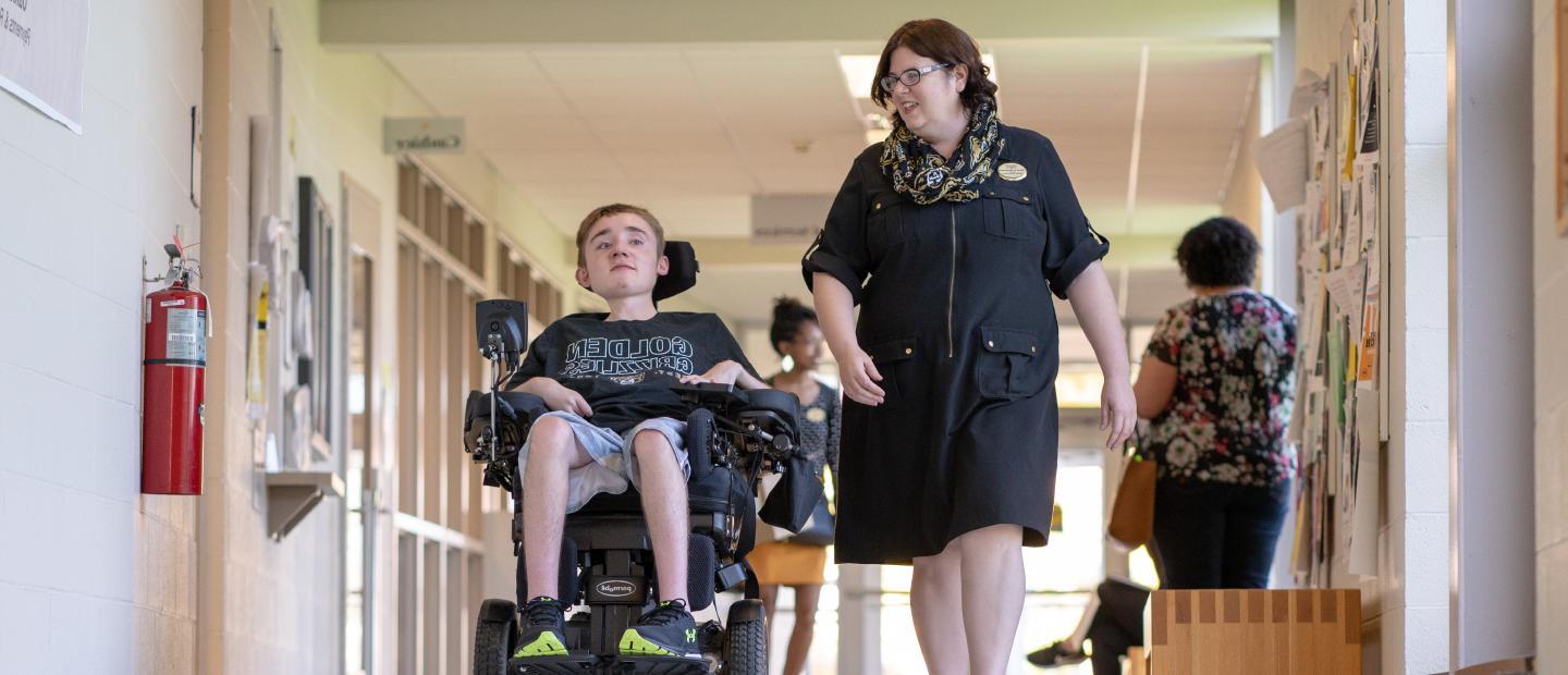一名女子走在走廊上，旁边坐着一个坐在轮椅上的年轻男子