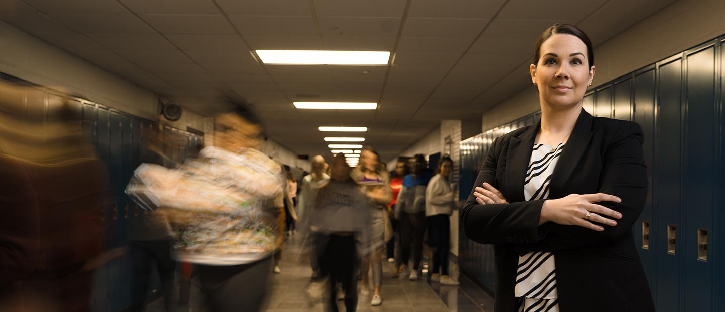 一名女子双手交叉站在储物柜走廊上，学生们走在她身后.